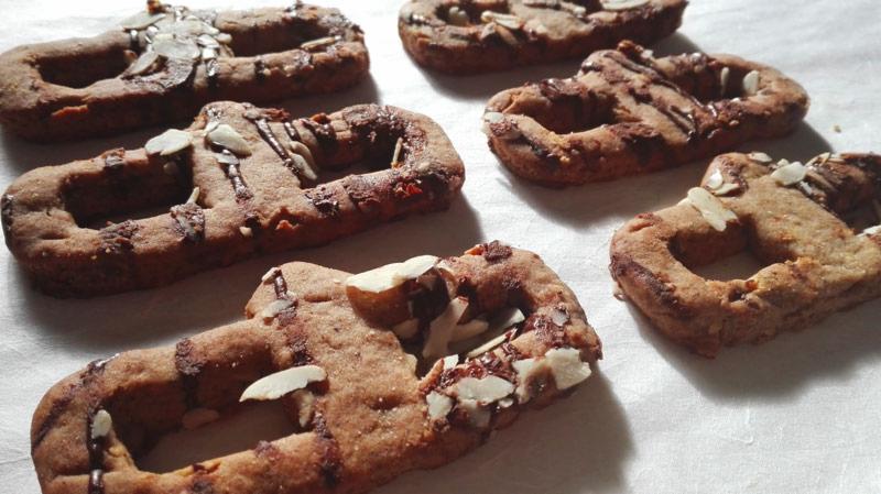 Ręcznie robione, brązowe ciasteczka w kształcie precli, udekorowane polewą czekoladową i płatkami migdałów, ułożone na białym tle.