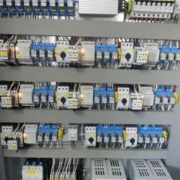 Szara szafa sterownicza z licznymi przekaźnikami, bezpiecznikami i przewodami, ułożonymi w rzędach, widoczne elementy z oznaczeniami Eaton oraz zasilacze impulsowe.