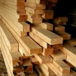 Stos jasnego, surowego drewna budowlanego ułożonego w nieładzie, widoczne przekroje belek i desek, w tle ciemna przestrzeń magazynowa.