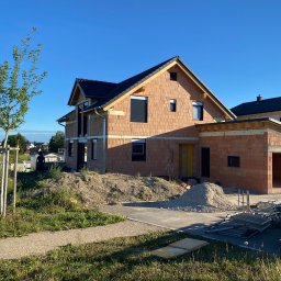 ZWARBAU - Dom w trakcie budowy z czerwonej cegły, widoczne rusztowania i materiały budowlane, błękitne niebo w tle. Stan surowy otwarty z garażem.