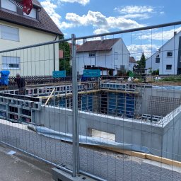 ZWARBAU - Wykopane fundamenty z betonową wylewką i szalunkiem na budowie domu jednorodzinnego. Widoczne zbrojenia i teren budowy zabezpieczony siatką.