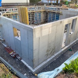 ZWARBAU - Surowy betonowy fundament domu z otworami okiennymi, widoczne szalunki i zbrojenia. Elementy konstrukcyjne w trakcie budowy, narzędzia w tle.