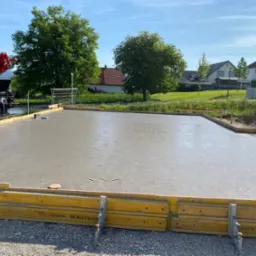Wylany fundament pod budowę domu, widoczne żółte szalunki. W tle drzewa i zabudowa jednorodzinna. Beton świeży, jeszcze mokry. Czerwony wysięgnik pompy do betonu.