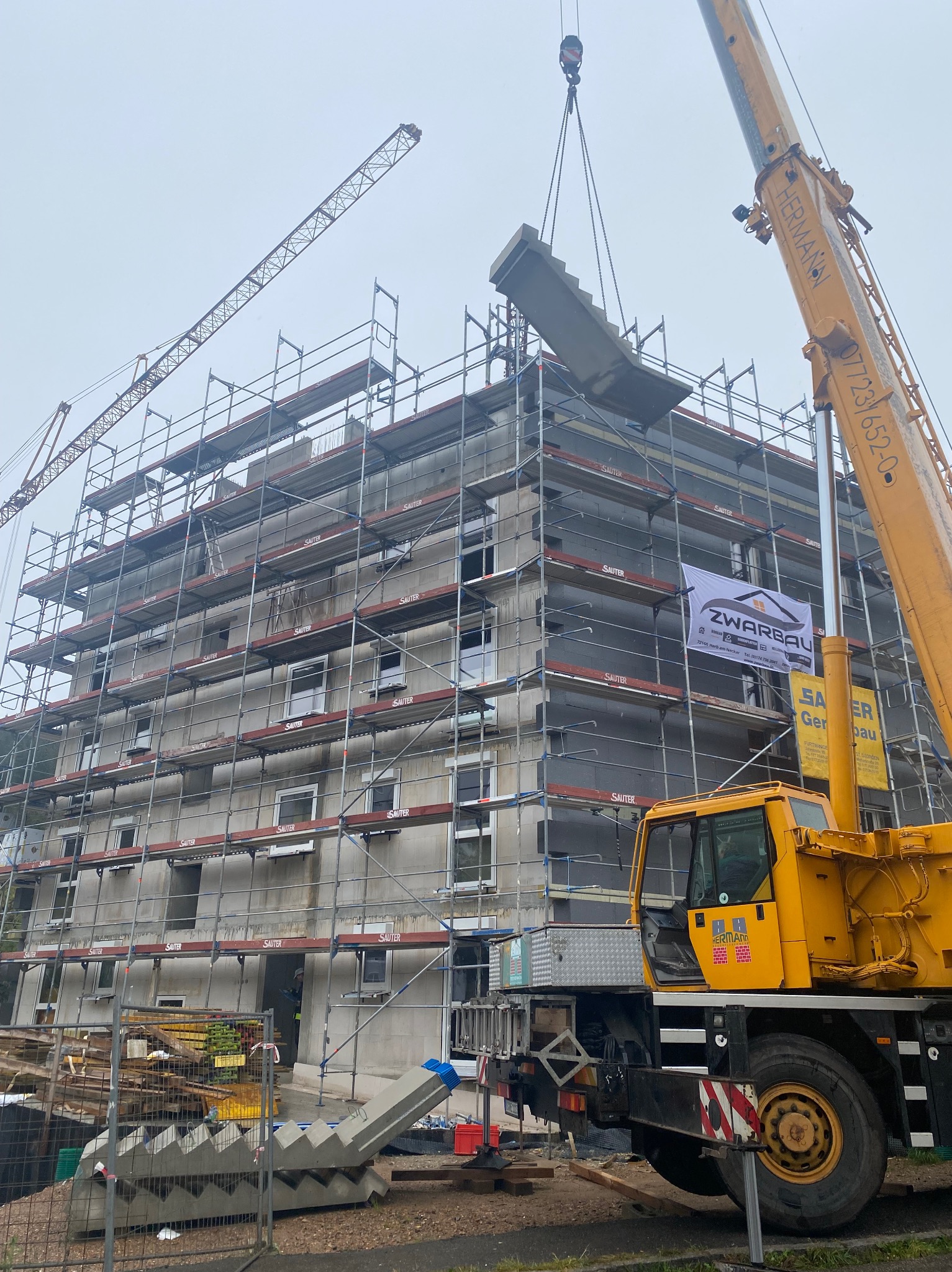 Montaż prefabrykowanych betonowych schodów zewnętrznych za pomocą dźwigu na tle budynku w rusztowaniach.