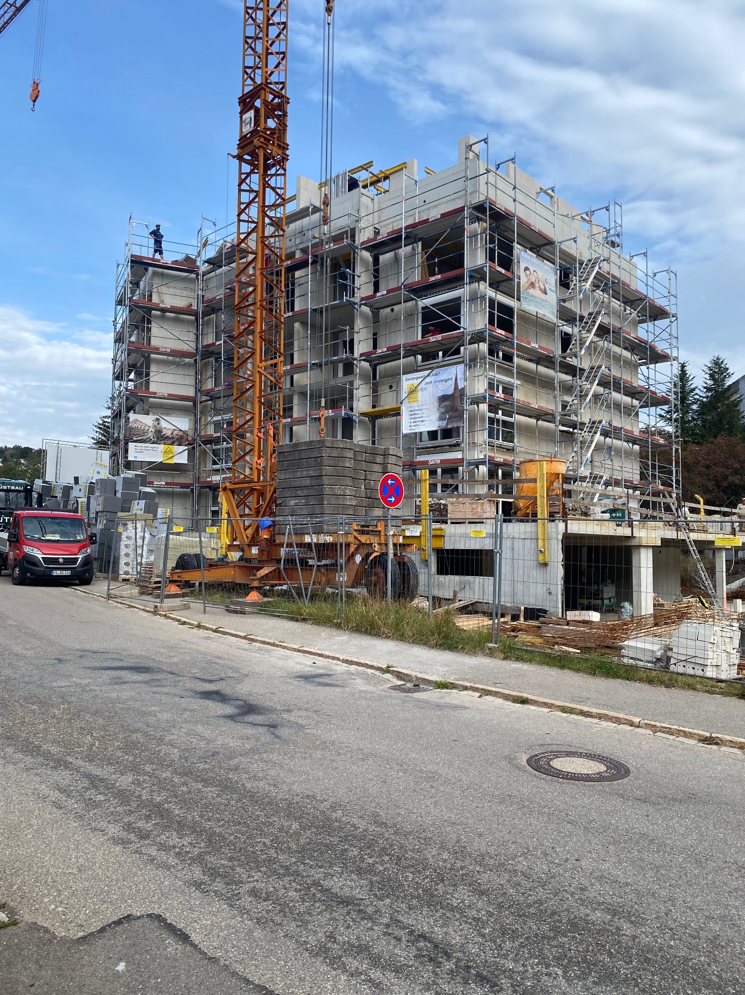 Wielopiętrowy budynek w trakcie budowy, otoczony rusztowaniami i z dźwigiem na pierwszym planie, widoczna ciężarówka z materiałami budowlanymi oraz robotnik na górze budynku.