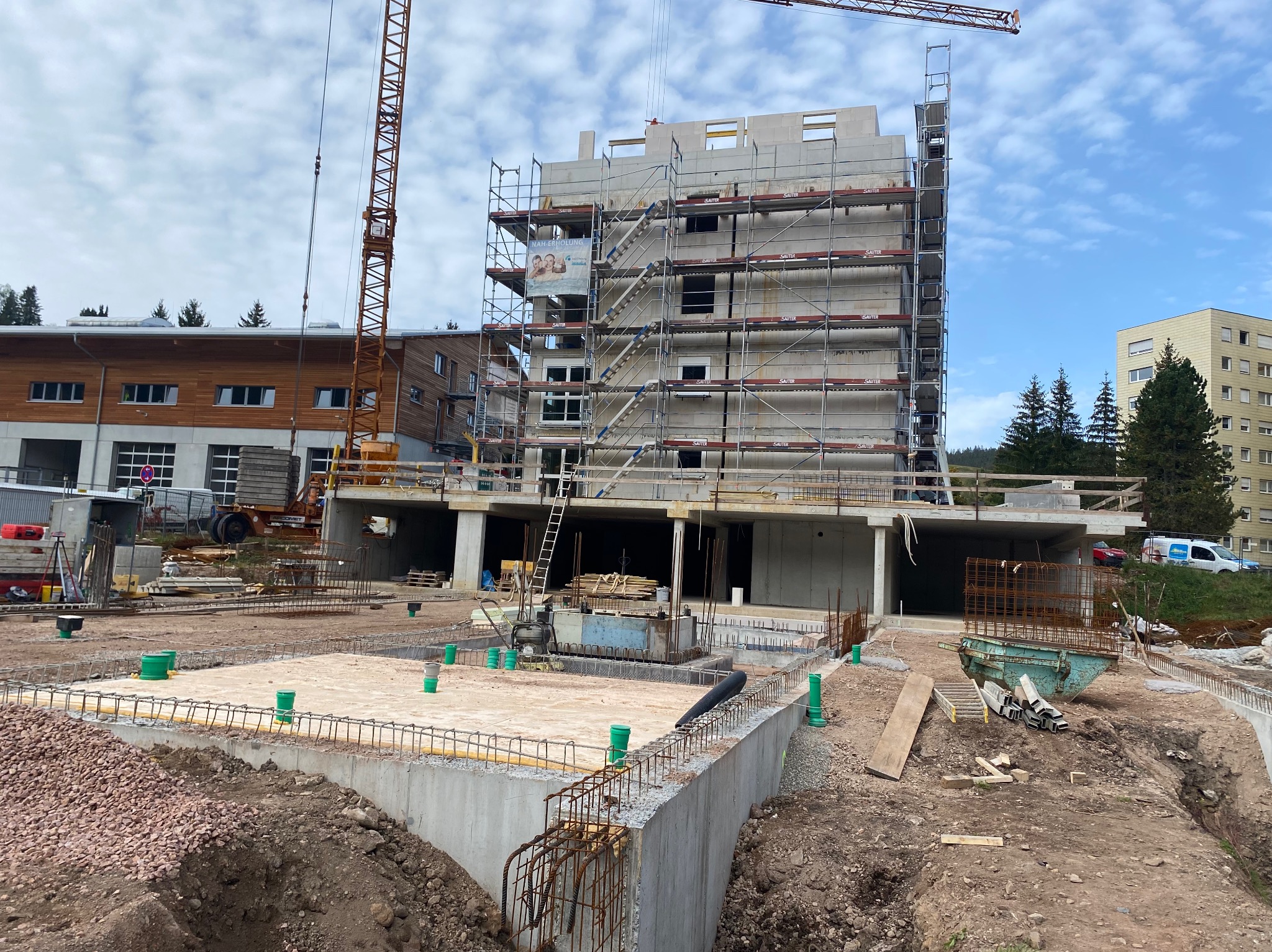 Wielopiętrowy budynek w trakcie budowy, otoczony rusztowaniami, z widocznymi fundamentami i konstrukcją żelbetową, na pierwszym planie wykop z wystającymi rurami wentylacyjnymi. Na budowie stoi...