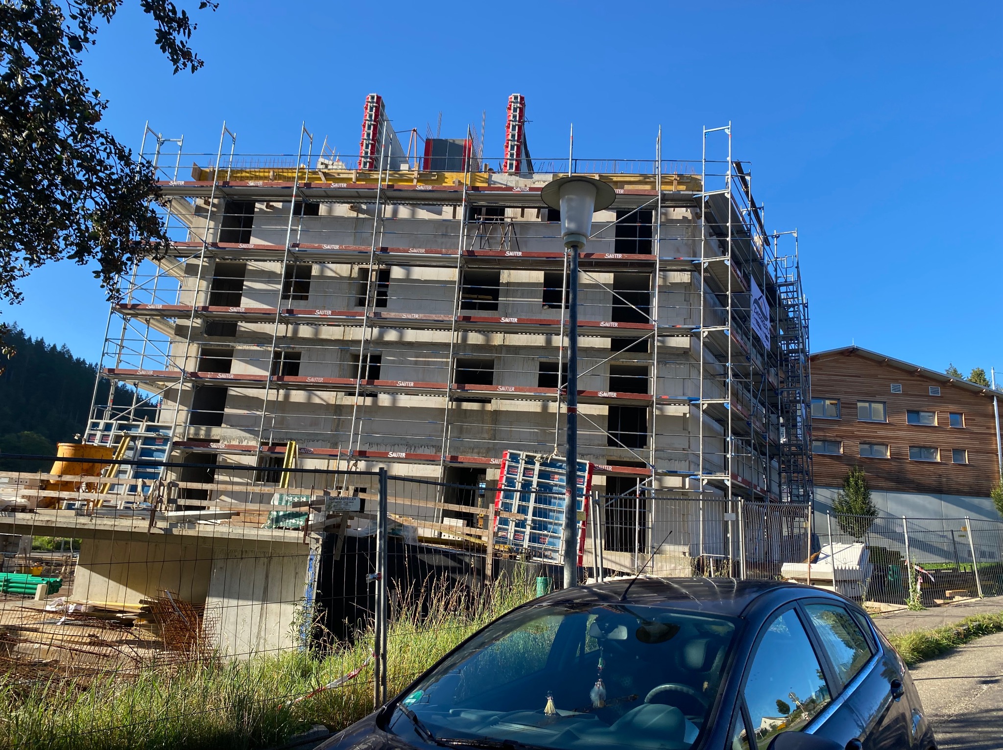 Surowy, czterokondygnacyjny budynek w trakcie budowy, otoczony rusztowaniami z logo Sauter, widoczny zza ogrodzenia i zaparkowanego samochodu na tle zieleni i błękitnego nieba.
