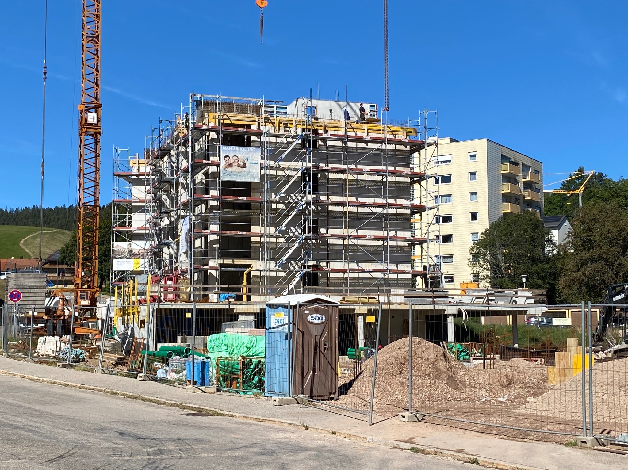 Wielopiętrowy budynek w trakcie budowy z rusztowaniami i dźwigiem, widoczne materiały budowlane i przenośna toaleta na pierwszym planie.