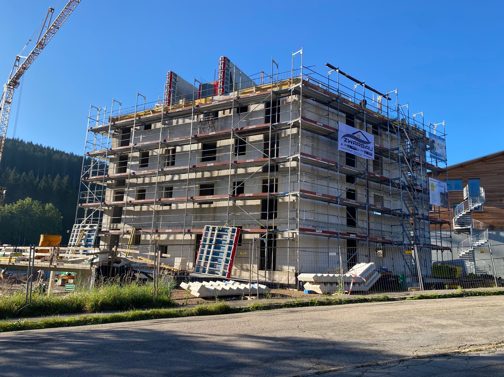Wielopiętrowy budynek w trakcie budowy, otoczony rusztowaniami, z widocznym żurawiem w tle i logo firmy ZWARBAU na banerze.