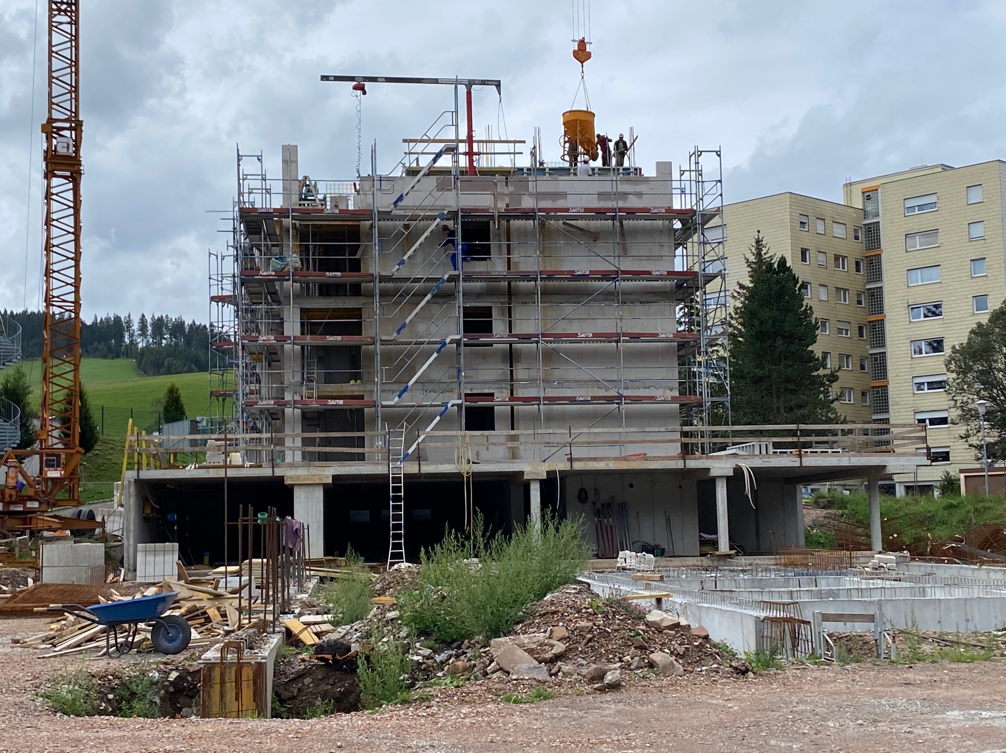 Budowa trzypiętrowego domu murowanego z betonu komórkowego, widoczne rusztowania, dźwig podający zaprawę i fundamenty, w tle budynek mieszkalny i zalesione wzgórze.