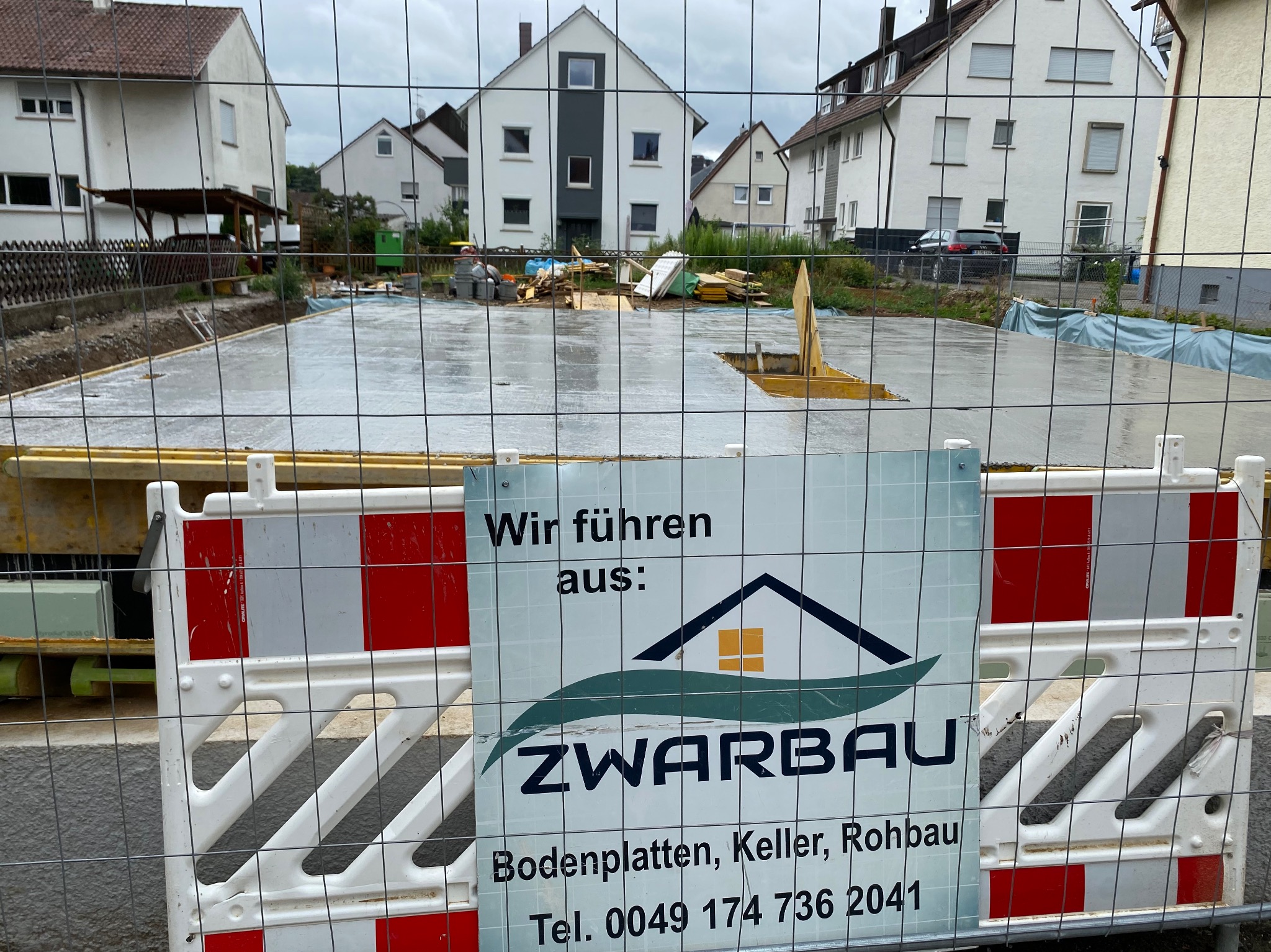 Wylany fundament pod budowę domu, zabezpieczony siatką i barierkami z tablicą informacyjną firmy ZWARBAU. W tle zabudowa jednorodzinna.