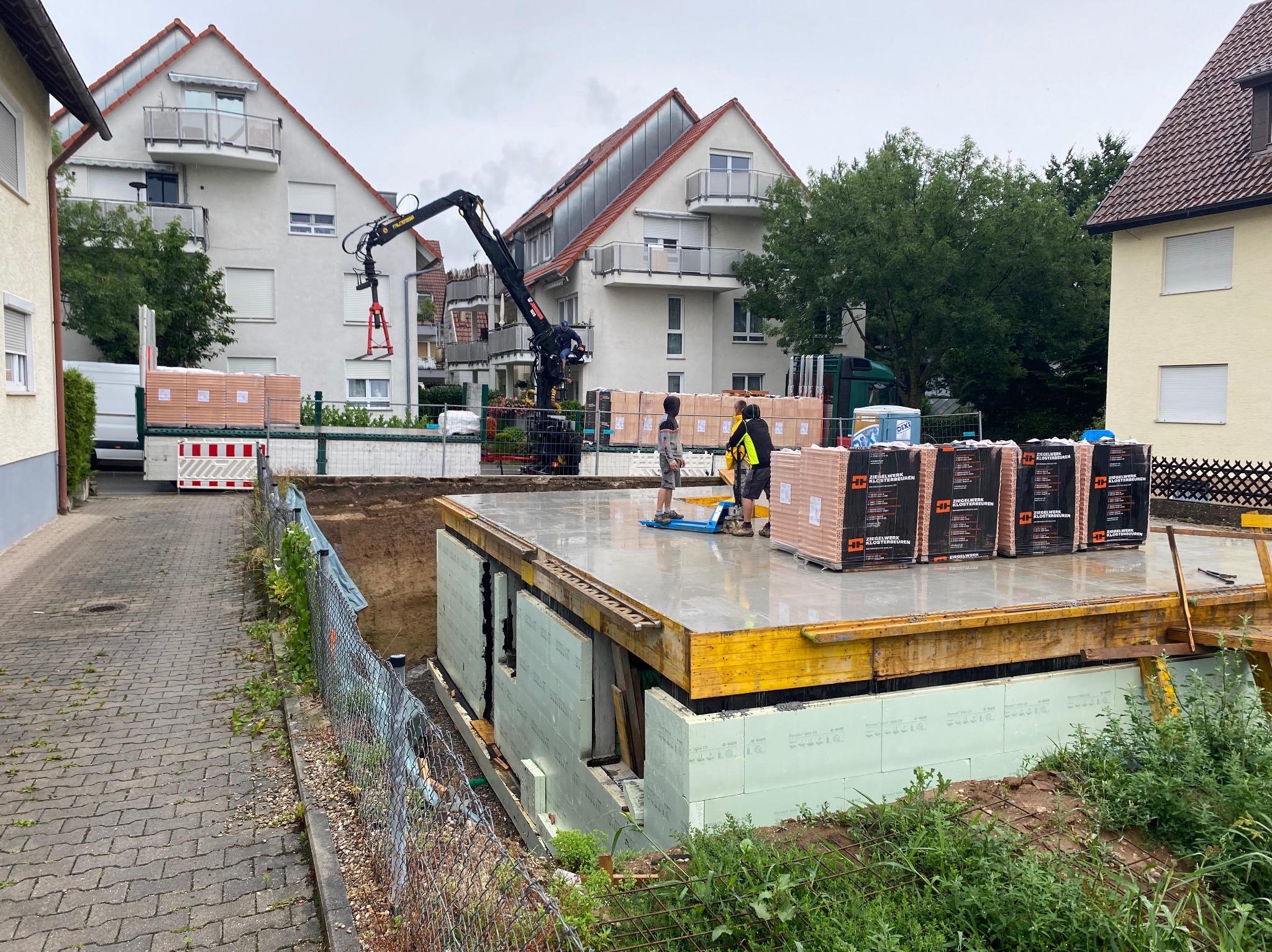 Budowa domu: fundamenty z hydroizolacją i termoizolacją, palety z cegłami na płycie fundamentowej, żuraw budowlany w tle. Etap wznoszenia ścian.