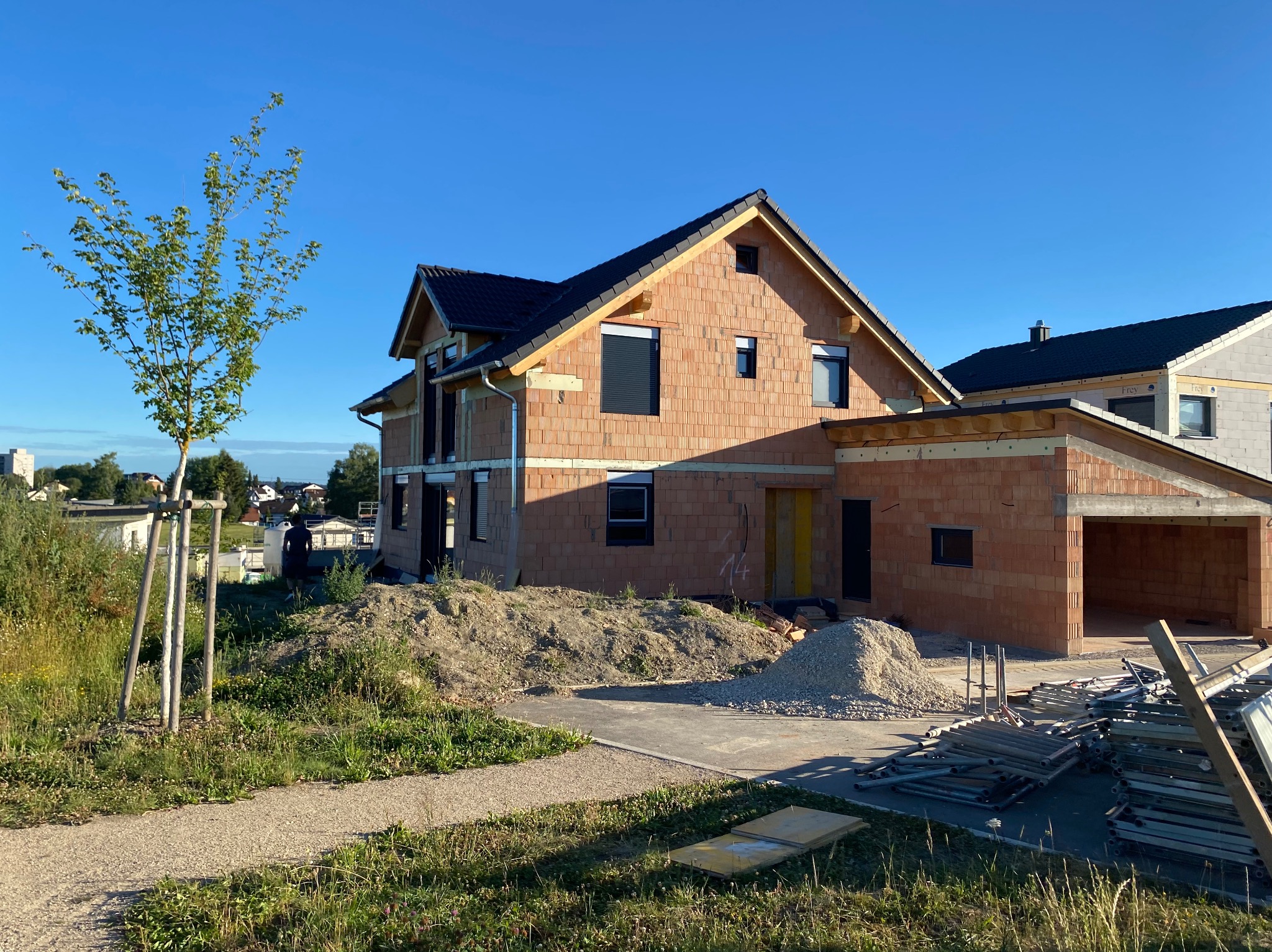 Dom w trakcie budowy z czerwonej cegły, widoczne rusztowania i materiały budowlane, błękitne niebo w tle. Stan surowy otwarty z garażem.