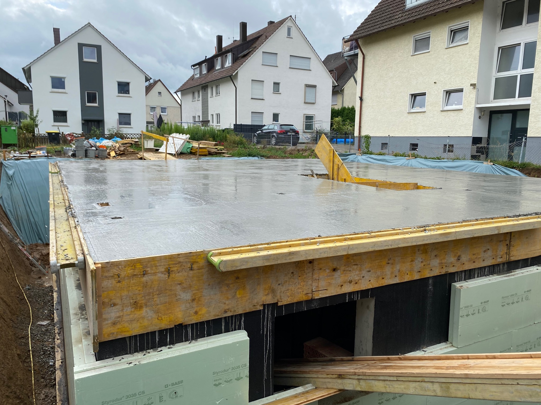 Zalane fundamenty domu z widocznym deskowaniem i izolacją termiczną. W tle zabudowa jednorodzinna i pochmurne niebo.