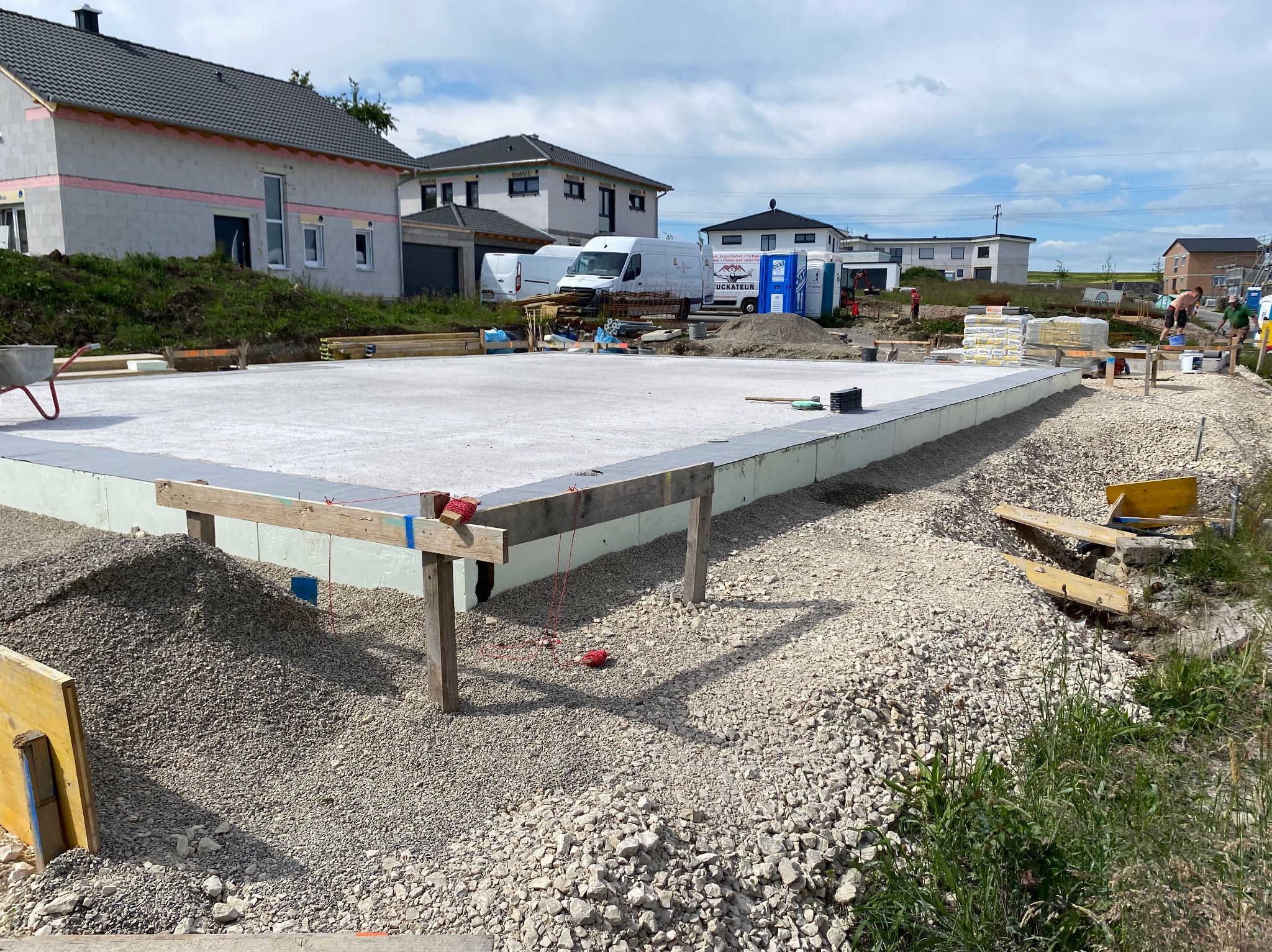 Wylany fundament pod budowę domu jednorodzinnego. Widoczne szalunki, izolacja termiczna i przygotowane podłoże z kruszywa. W tle zabudowa mieszkalna i błękitne niebo.