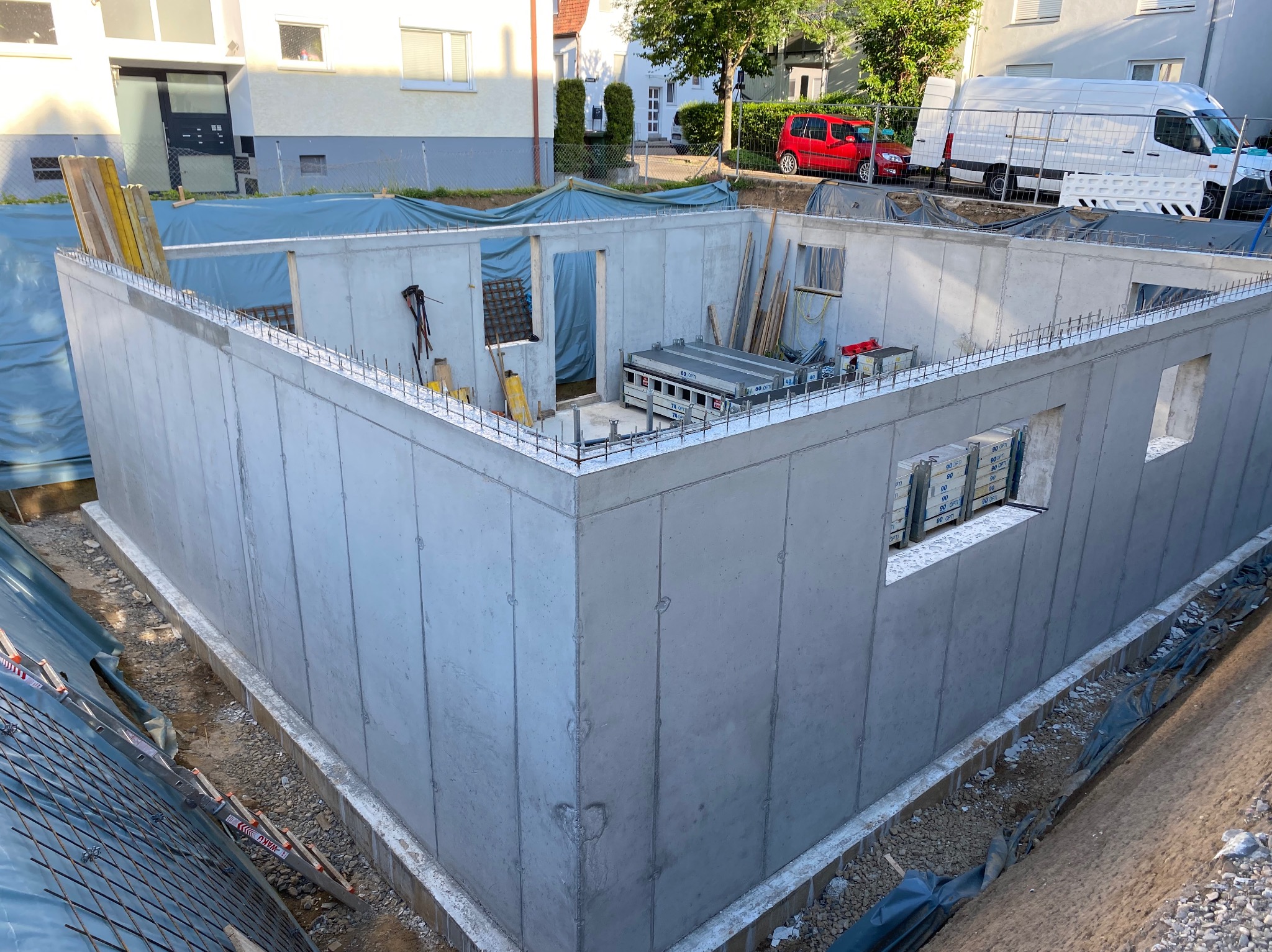 Surowy, betonowy fundament domu z otworami okiennymi i zbrojeniem, otoczony wykopem. Widoczne narzędzia i materiały budowlane. Tło: budynki mieszkalne i samochody.