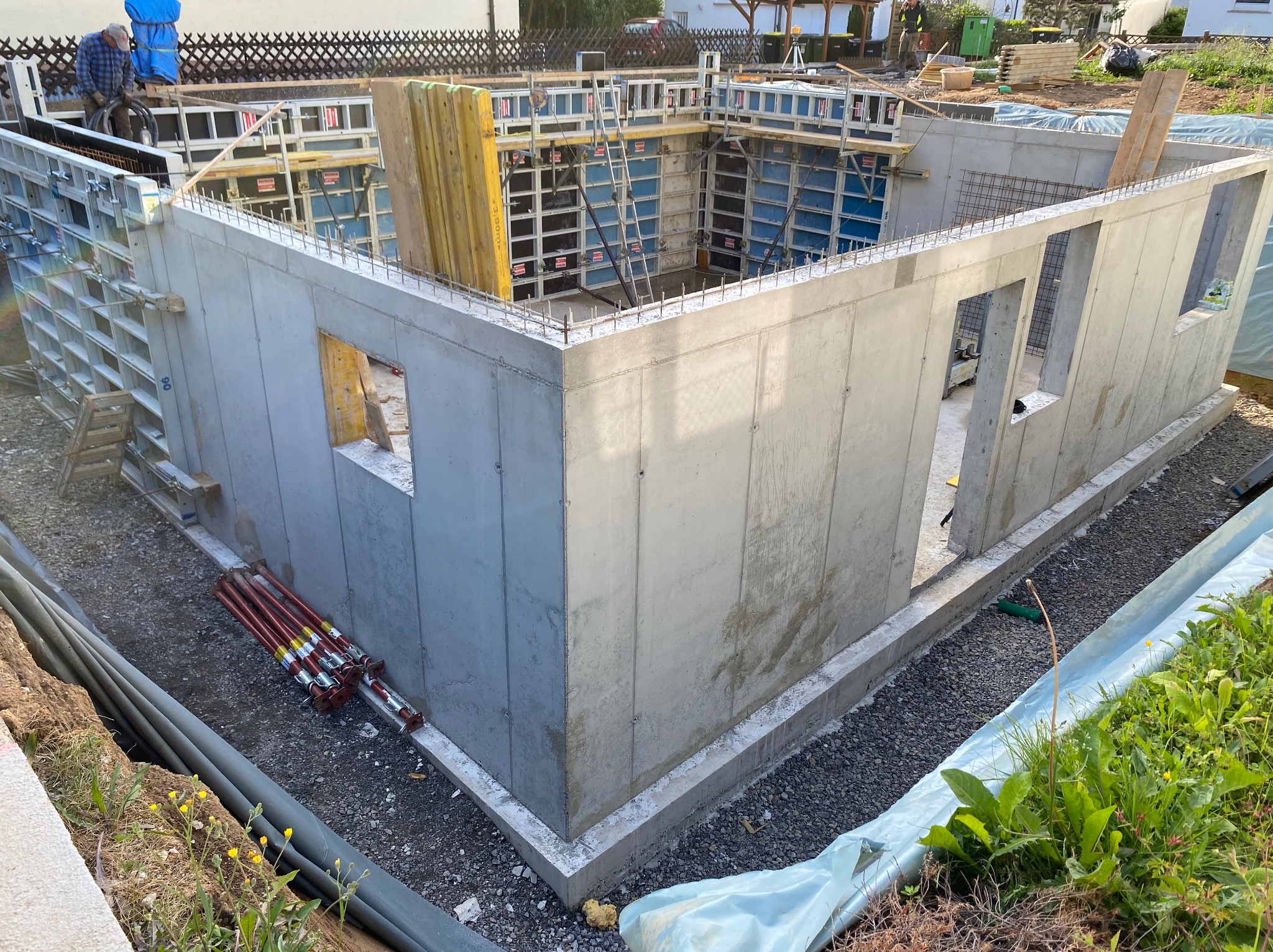 Surowy betonowy fundament domu z otworami okiennymi, widoczne szalunki i zbrojenia. Elementy konstrukcyjne w trakcie budowy, narzędzia w tle.