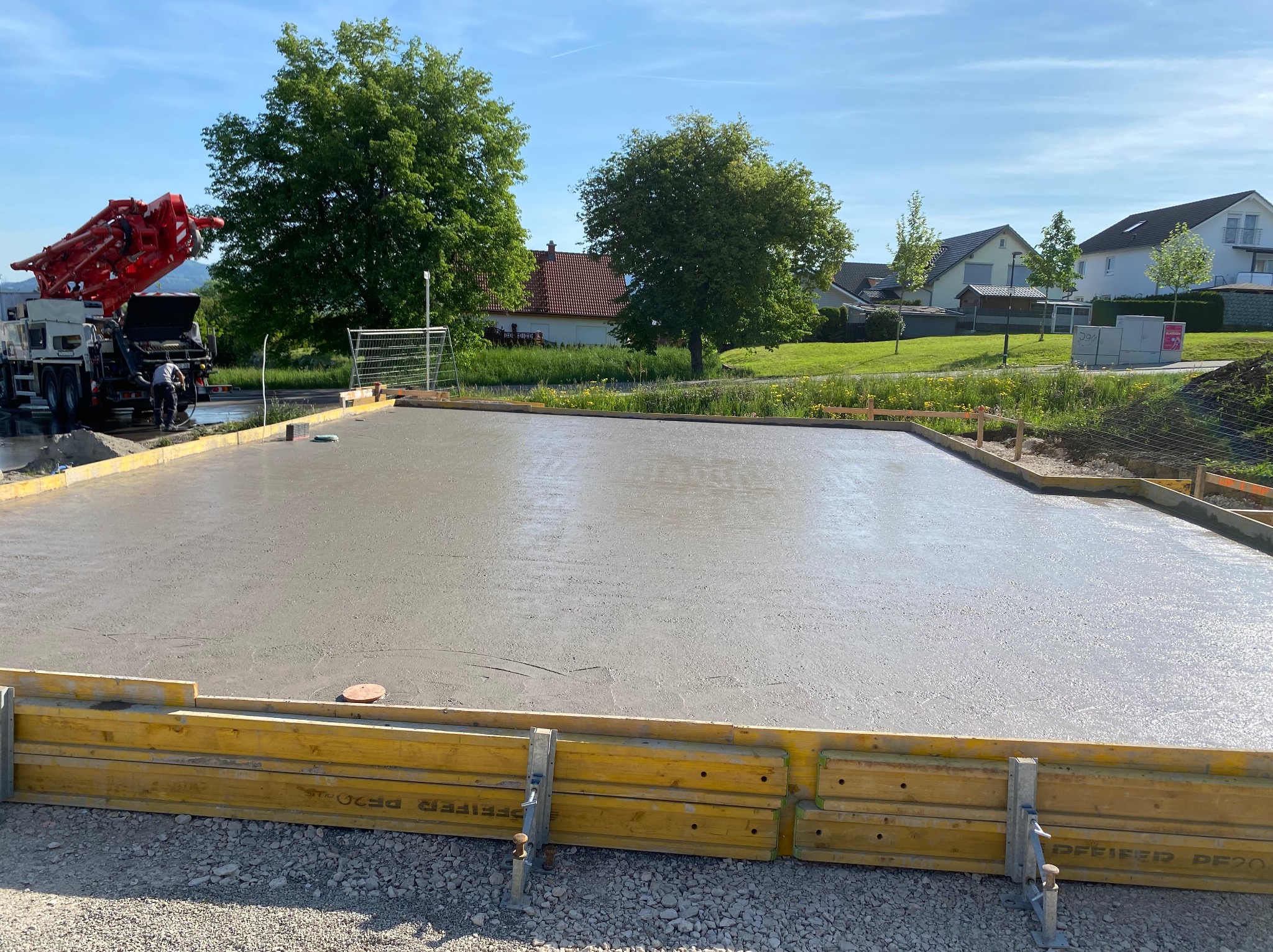 Wylany fundament pod budowę domu, widoczne żółte szalunki. W tle drzewa i zabudowa jednorodzinna. Beton świeży, jeszcze mokry. Czerwony wysięgnik pompy do betonu.