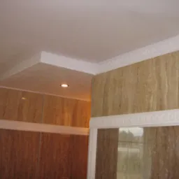 Wykończona łazienka z beżowymi płytkami imitującymi marmur, ozdobnymi białymi listwami sufitowymi i oświetleniem punktowym. Widok na narożnik pomieszczenia.