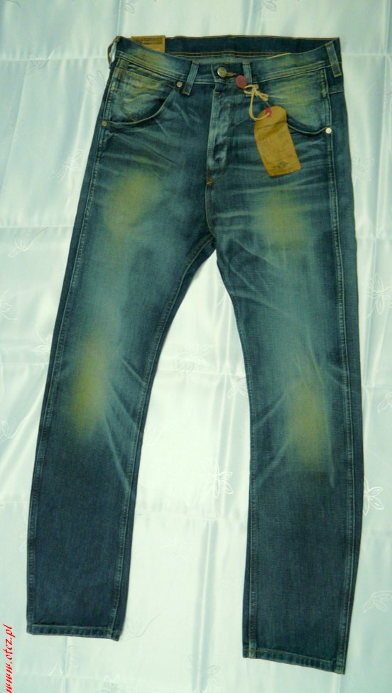 Rozłożone dżinsy Wrangler Lee w odcieniach niebieskiego z efektem sprania i brązową papierową metką na tle białej tkaniny z delikatnym wzorem.