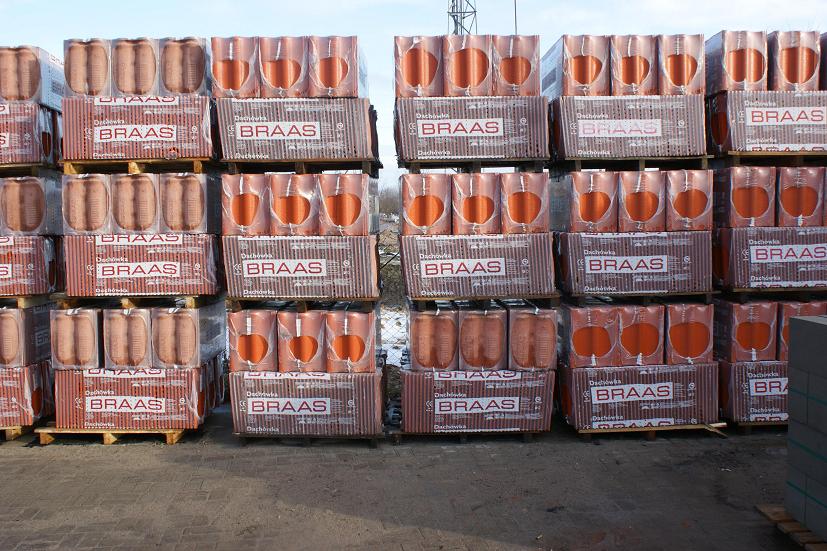 Palety z czerwonymi dachówkami betonowymi marki Braas, zapakowane w folię, składowane na placu budowy, widoczne logo producenta na opakowaniach.