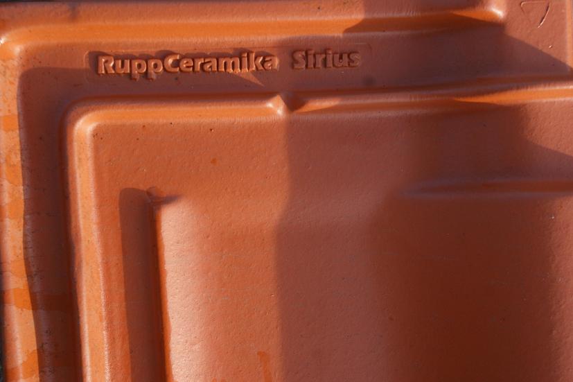 Ceglasta dachówka ceramiczna marki Rupp Ceramika Sirius, z widocznym logo producenta, oświetlona bocznym światłem.