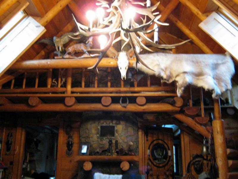 Wnętrze drewnianego domu z wysokim stropem i dekoracyjnym żyrandolem z poroża. Widoczny kamienny kominek i trofea myśliwskie.