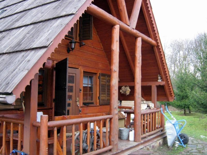 Drewniany dom z gontowym dachem, widok na werandę z balustradą i otwarte drzwi. Charakterystyczne zdobienia okapów i drewniane kolumny. W tle zieleń drzew.