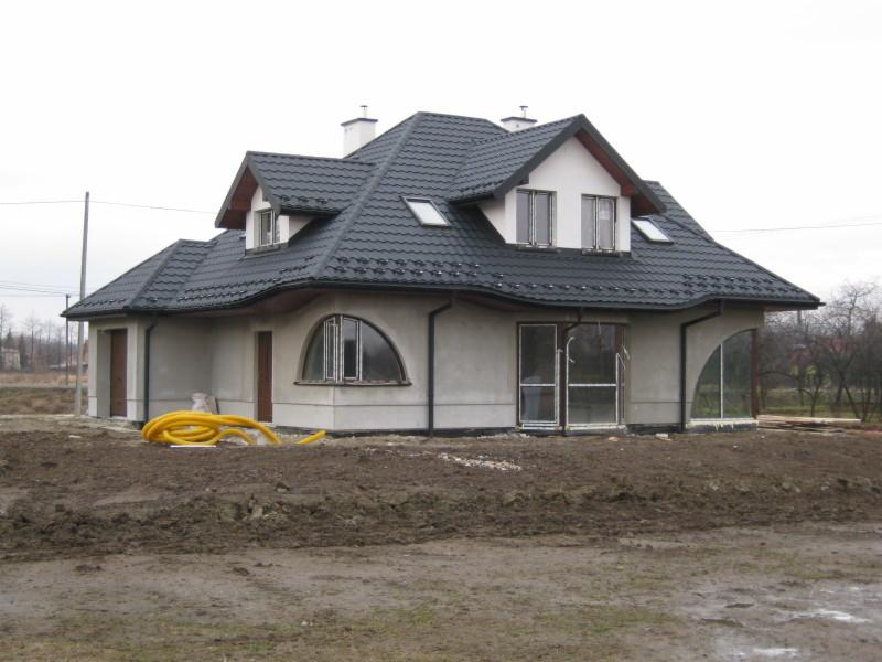 Dom jednorodzinny z ciemnym dachem krytym dachówką, widoczne okna dachowe i rynny. Na pierwszym planie ziemia i żółte rury. Budynek w stanie surowym.