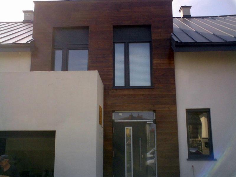 Elewacja domu z drewnianą okładziną i szarym dachem. Widoczne okna z roletami i nowoczesne drzwi wejściowe. Uwagę zwraca kontrast materiałów i kolorów.