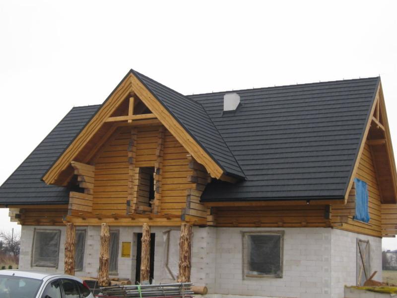 Surowy stan domu z bali z ciemnym dachem z dachówki, widoczne drewniane elementy konstrukcyjne i białe bloczki fundamentowe. Częściowo zakryte okna folią.