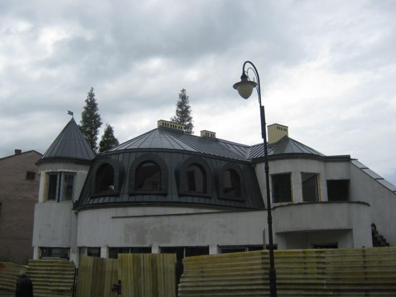 Budynek z nietypowym, okrągłym dachem pokrytym ciemną blachą, z wieżyczką i lukarnami w kształcie łuków. W tle pochmurne niebo i drzewa.