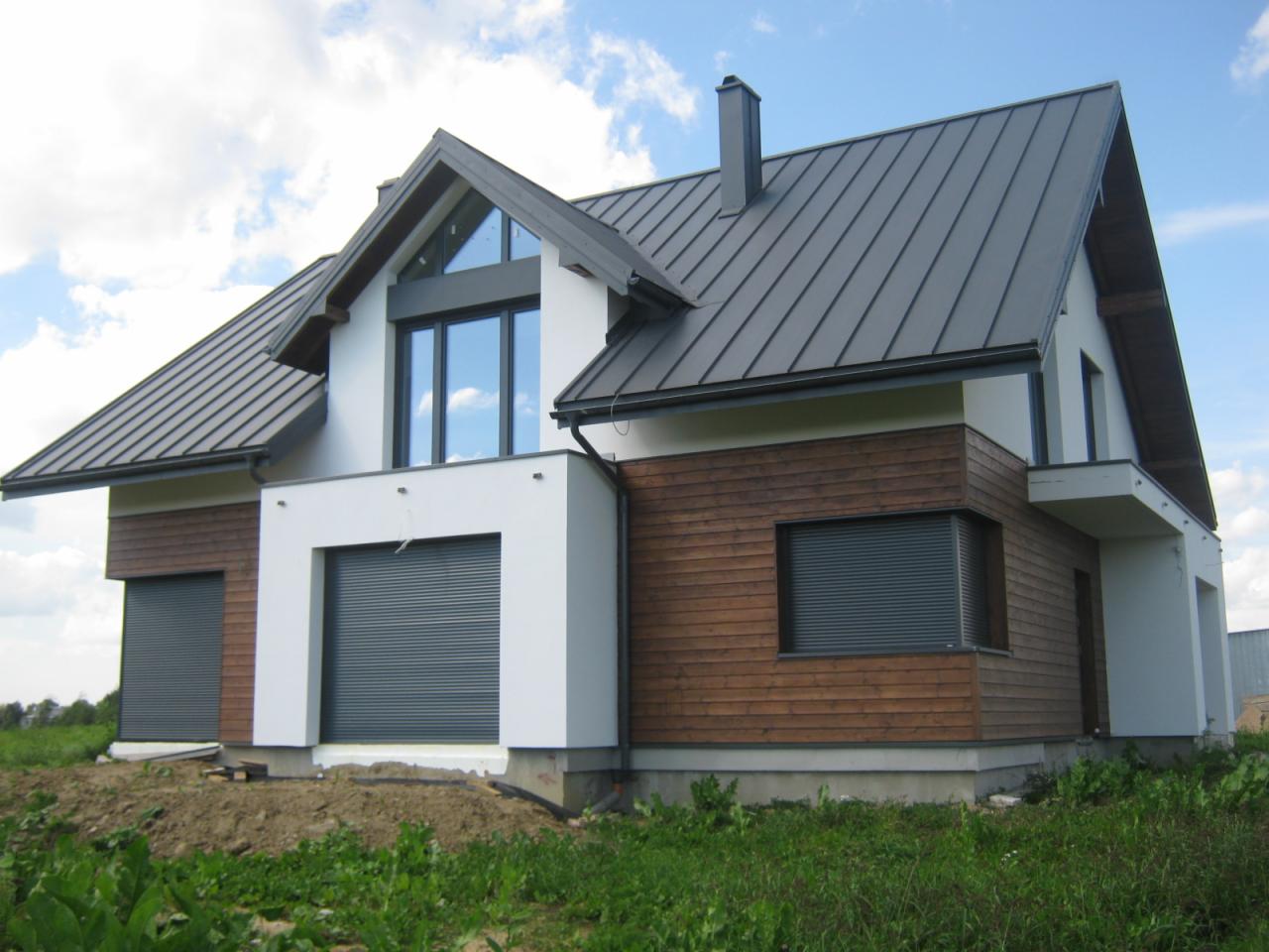 Nowoczesny dom jednorodzinny z ciemnoszarą blachodachówką, drewnianą elewacją i roletami zewnętrznymi. Widoczne okna z refleksami nieba i białe elementy architektoniczne.