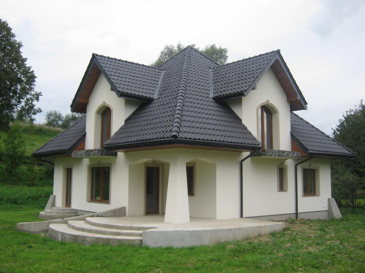 Dom z ciemną dachówką ceramiczną, jasną elewacją i drewnianymi elementami wykończeniowymi. Widoczne schody wejściowe i zadbany trawnik. Ujęcie z poziomu gruntu.