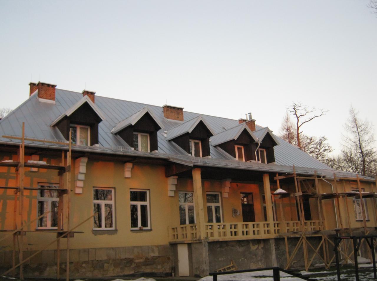 Budynek z szarym dachem i lukarnami, w trakcie prac remontowych. Widoczne rusztowania, jasna elewacja i fragmenty ośnieżonego terenu. Ujęcie za dnia.