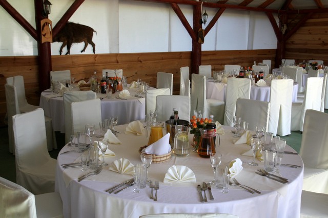 Sala bankietowa z okrągłymi stołami nakrytymi białymi obrusami i krzesłami w białych pokrowcach, na ścianie drewniana płaskorzeźba żubra, przygotowana na imprezę.