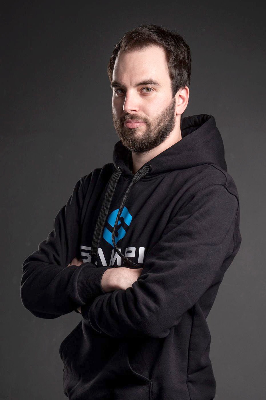 Mężczyzna w czarnej bluzie z kapturem i nadrukiem logo na piersi, pozujący na szarym tle ze skrzyżowanymi rękami. Portret w półzbliżeniu.