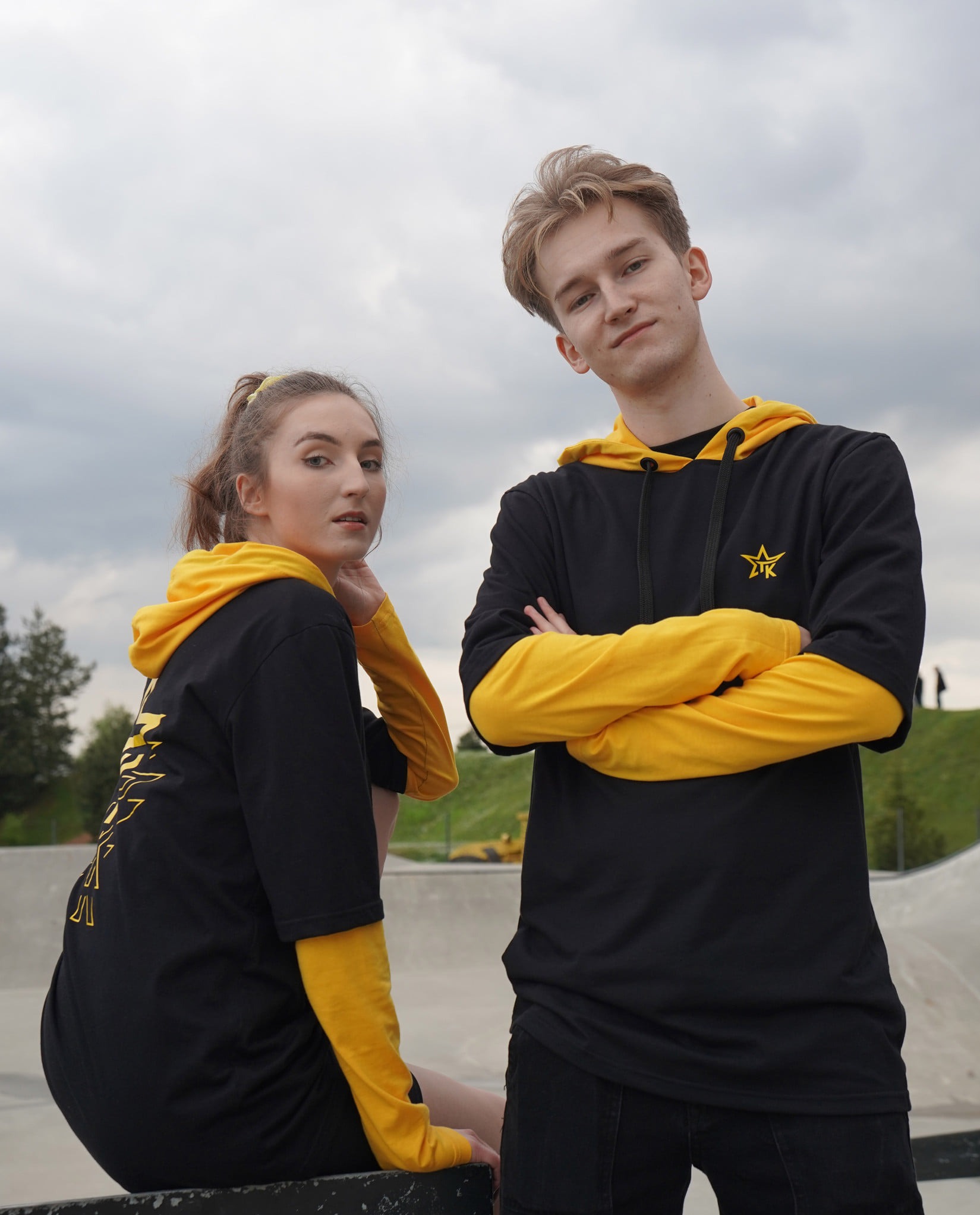 Dwoje młodych ludzi prezentuje czarno-żółte bluzy z kapturem i nadrukiem gwiazdy na tle skateparku. Ujęcie z dołu podkreśla młodzieżowy styl odzieży.