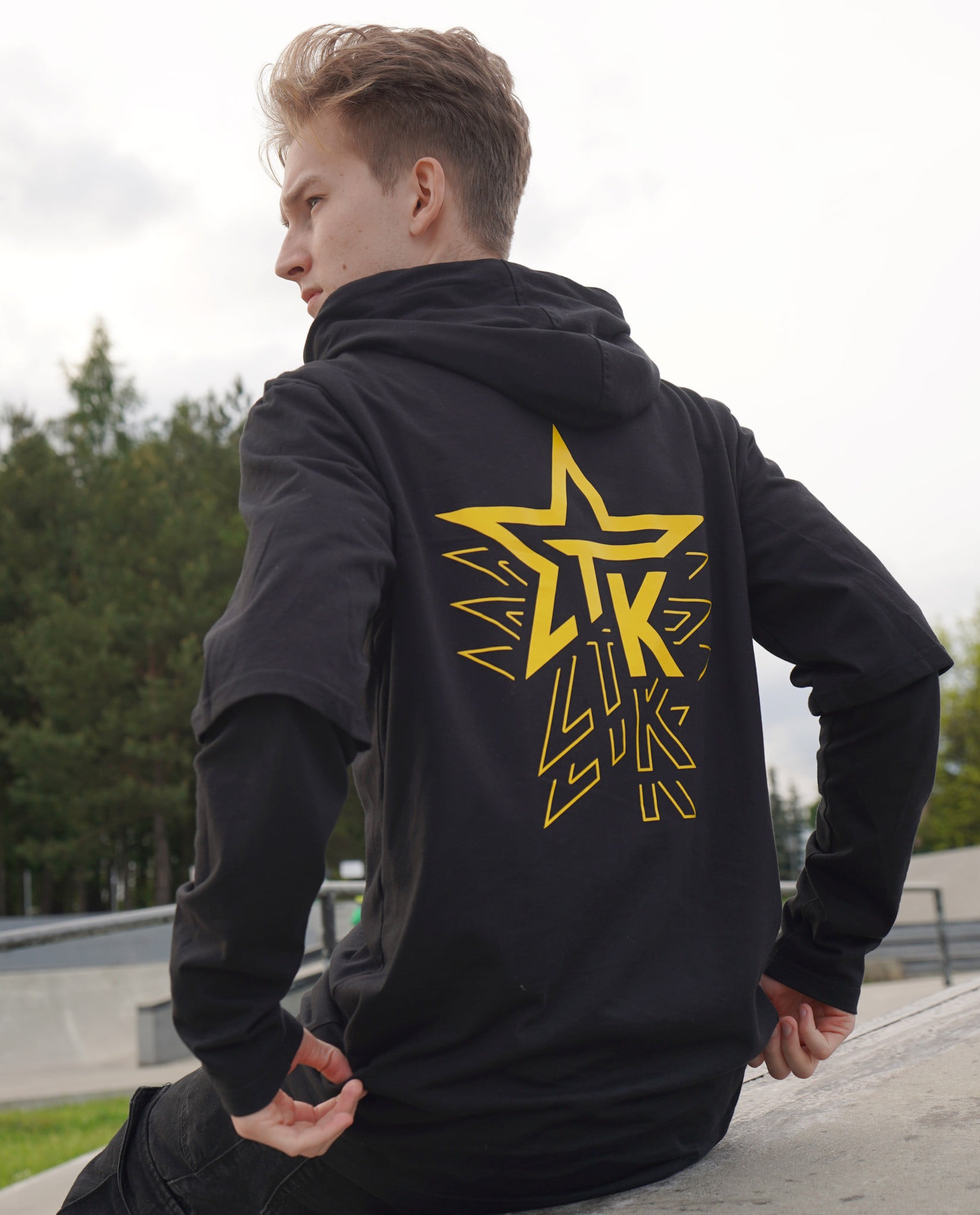 Młodzieniec w czarnej bluzie z kapturem, na której plecach widnieje żółty nadruk gwiazdy i napisu. Ujęcie z tyłu, skatepark w tle.