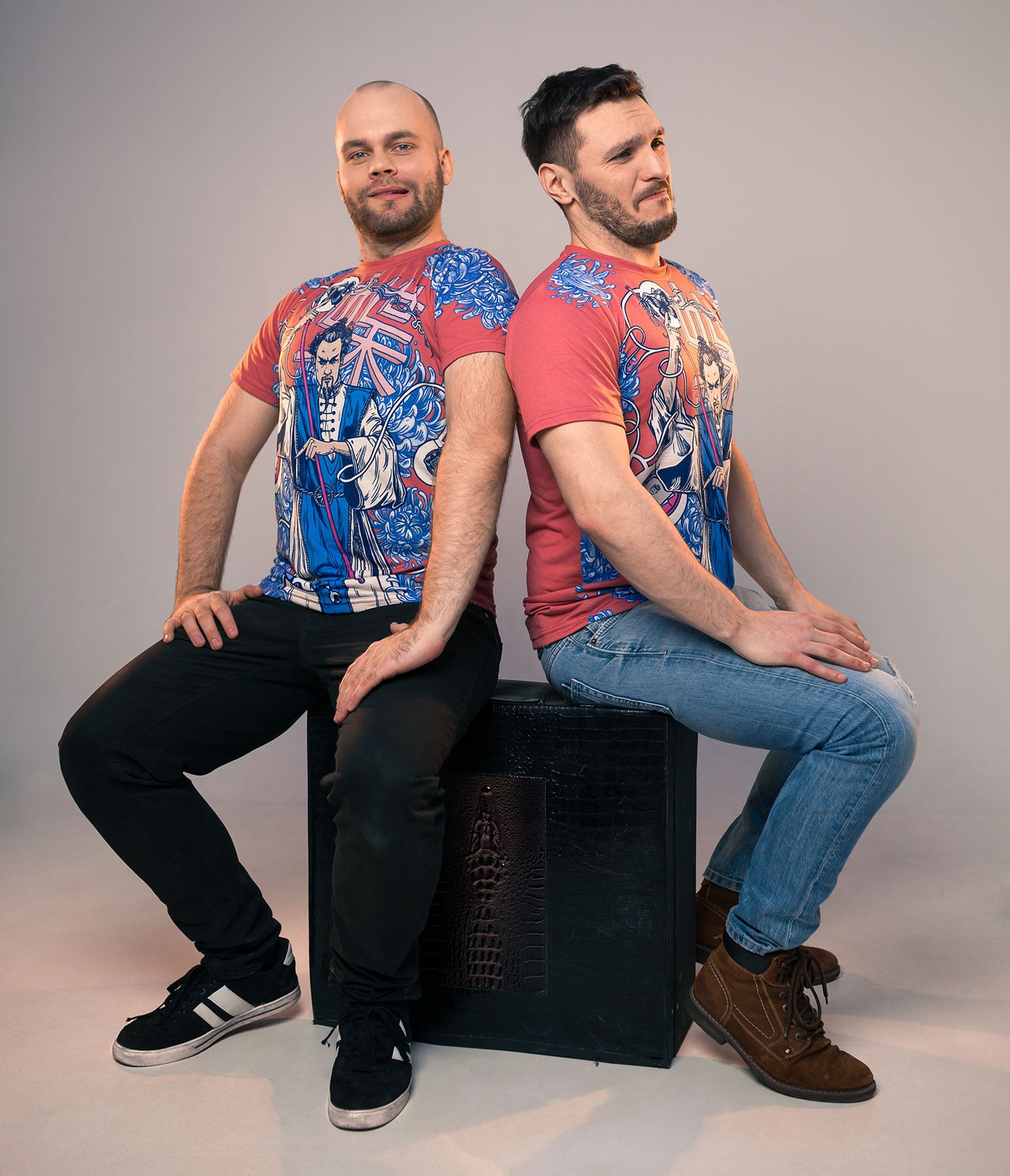 Dwóch mężczyzn siedzi tyłem do siebie na czarnym podeście, prezentując koszulki z unikalnym nadrukiem w stylu azjatyckim na czerwonym tle.