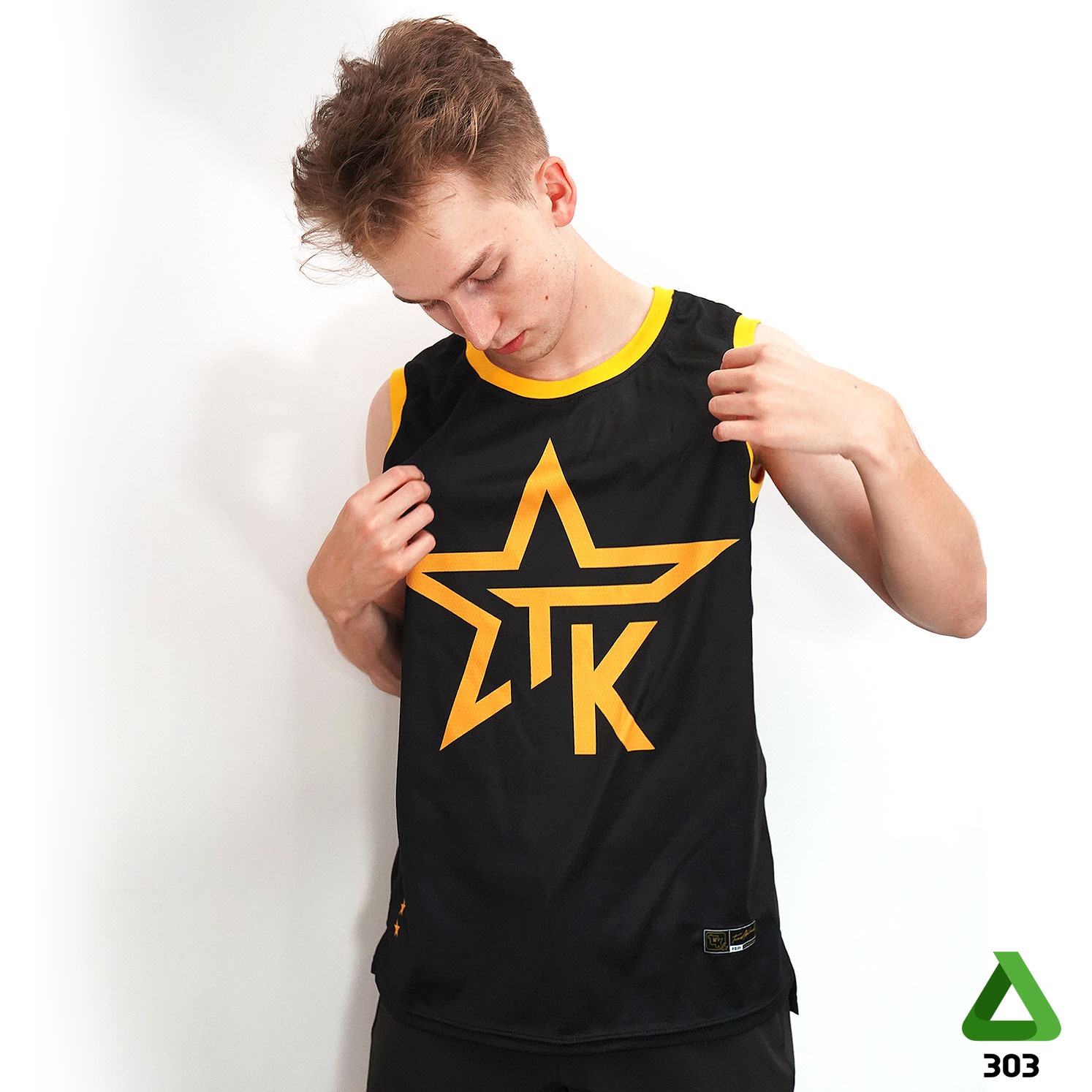 Młody mężczyzna prezentuje czarną koszulkę sportową bez rękawów z żółtym nadrukiem gwiazdy i liter, stojąc na tle białej ściany.