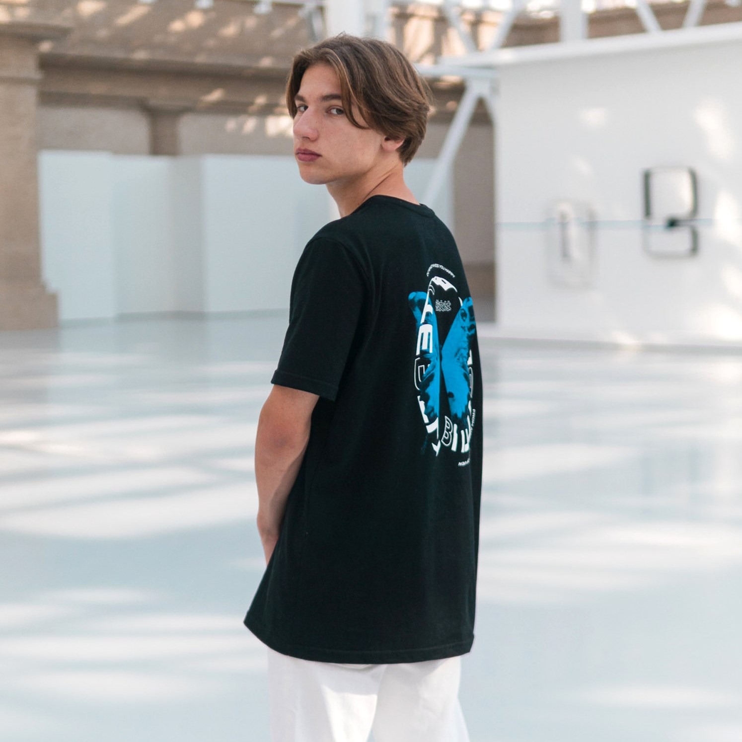 Młody mężczyzna w czarnej koszulce z nadrukiem motyla na plecach, odwrócony tyłem, w jasnym, minimalistycznym wnętrzu. Prezentacja nadruku na odzieży.