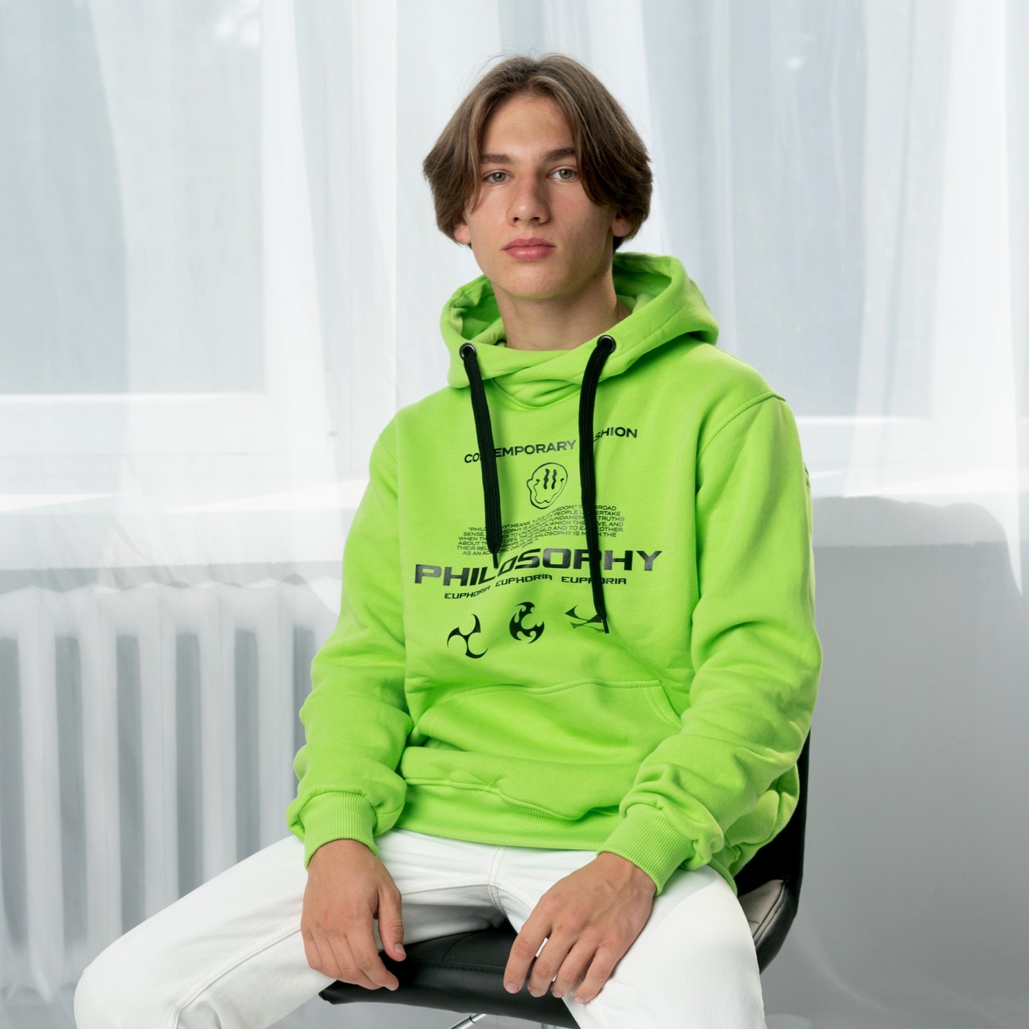 Młody mężczyzna siedzi na krześle, prezentując jasnozieloną bluzę z kapturem i nadrukiem 'Philosophy'. Białe spodnie i stonowane tło podkreślają wyrazistość bluzy.