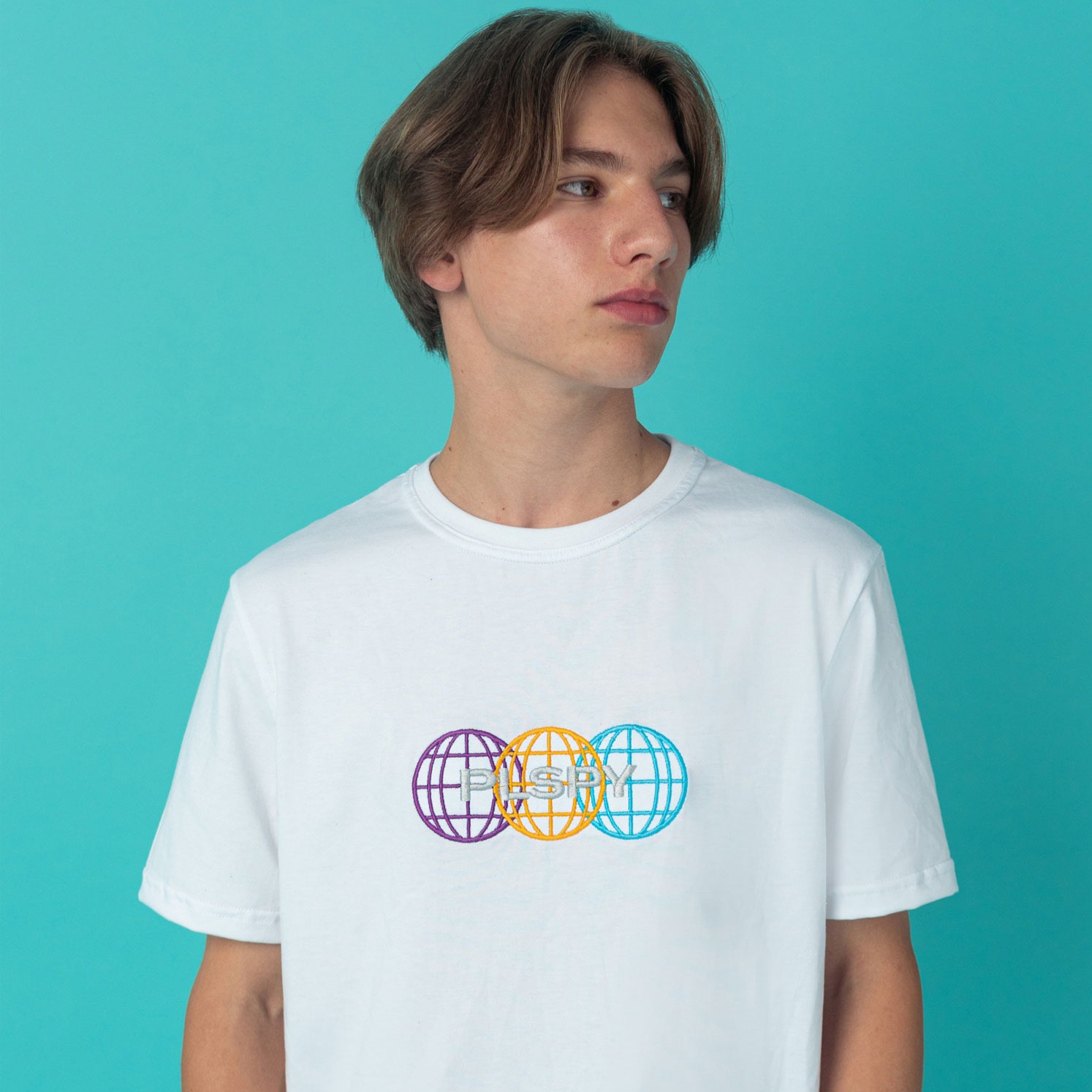 Młody mężczyzna prezentuje białą koszulkę z kolorowym, graficznym nadrukiem trzech globusów z napisem 'PLSPY' na tle turkusowego studia.