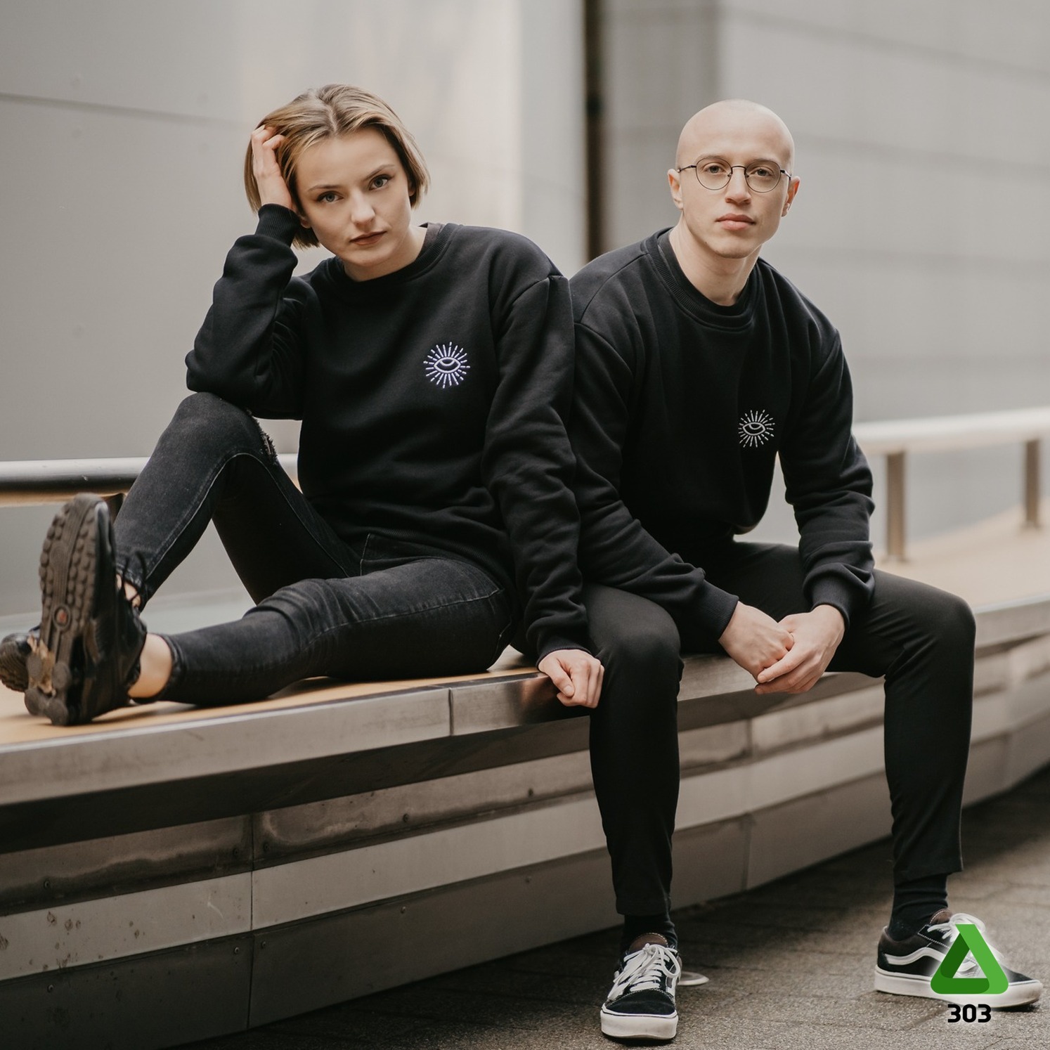 Dwoje modeli prezentuje czarne bluzy z unikalnym, białym nadrukiem oka na piersi, siedząc na metalowej ławce na tle szarej ściany. Stonowana kolorystyka, miejski styl.