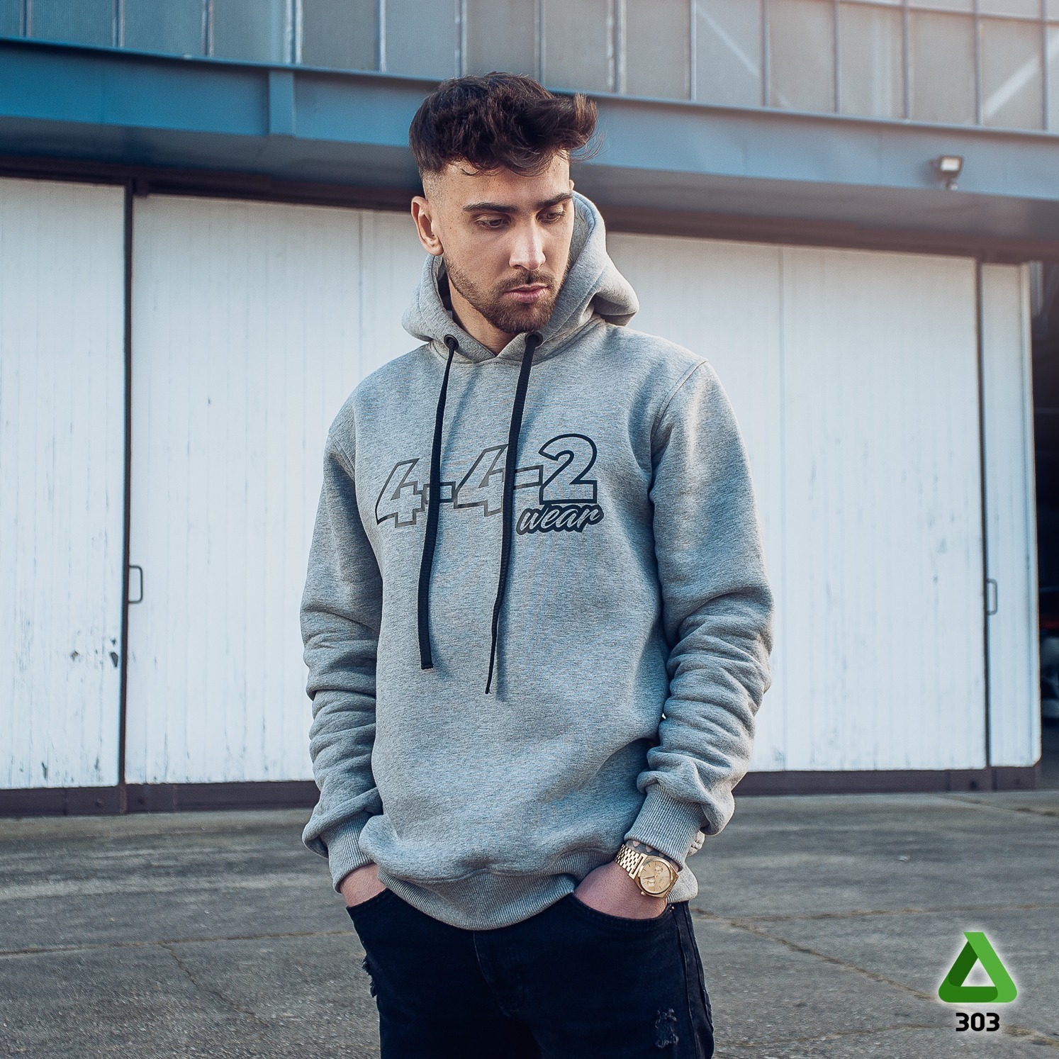 Mężczyzna w szarej bluzie z kapturem i czarnym nadrukiem '4-4-2 wear', pozuje na tle białych drzwi hangaru. Widoczny złoty zegarek i podarte jeansy.