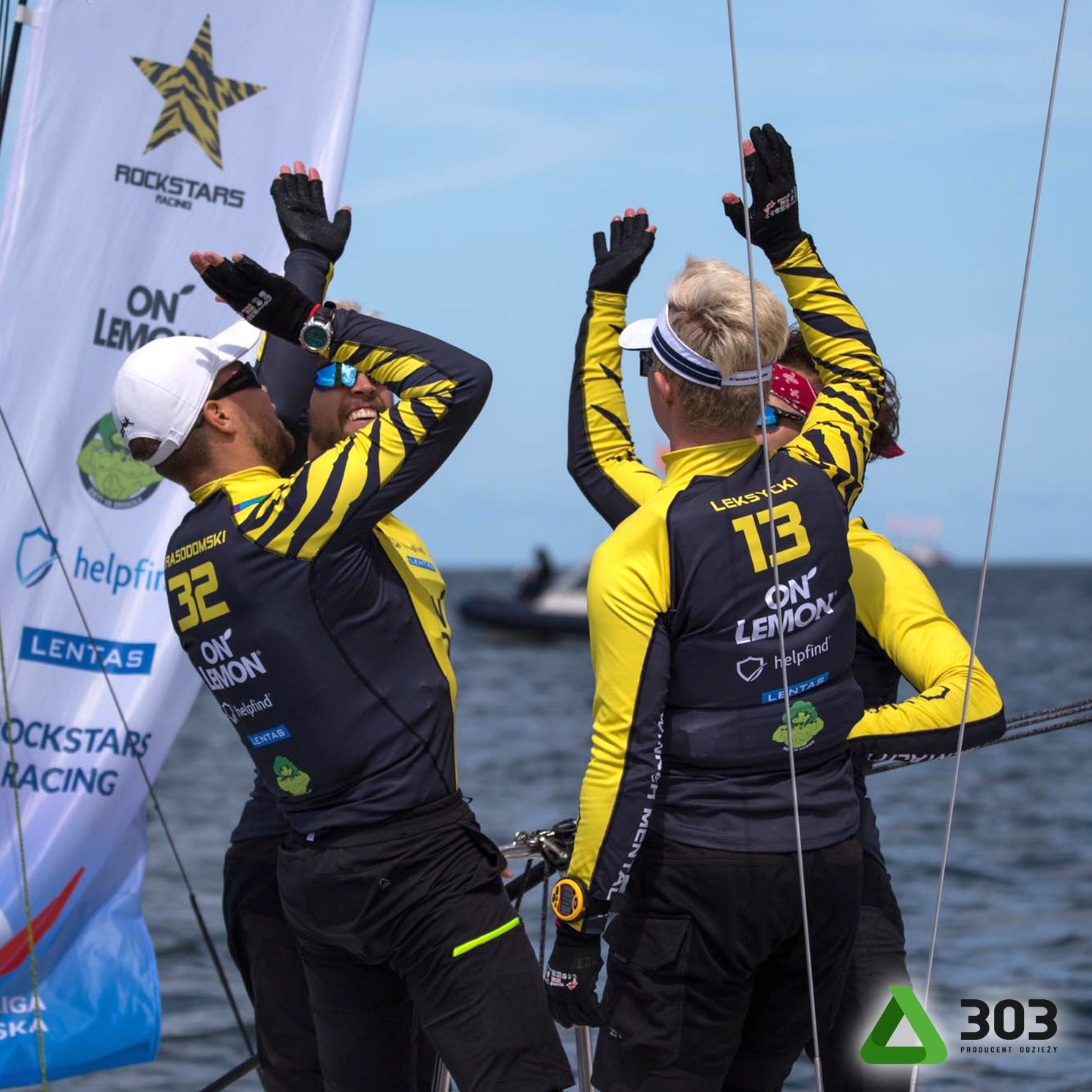 Załoga jachtu w spersonalizowanych, czarno-żółtych strojach z nadrukami, świętuje sukces na tle morza i flagi sponsorskiej. Widoczne numery i nazwy zawodników.