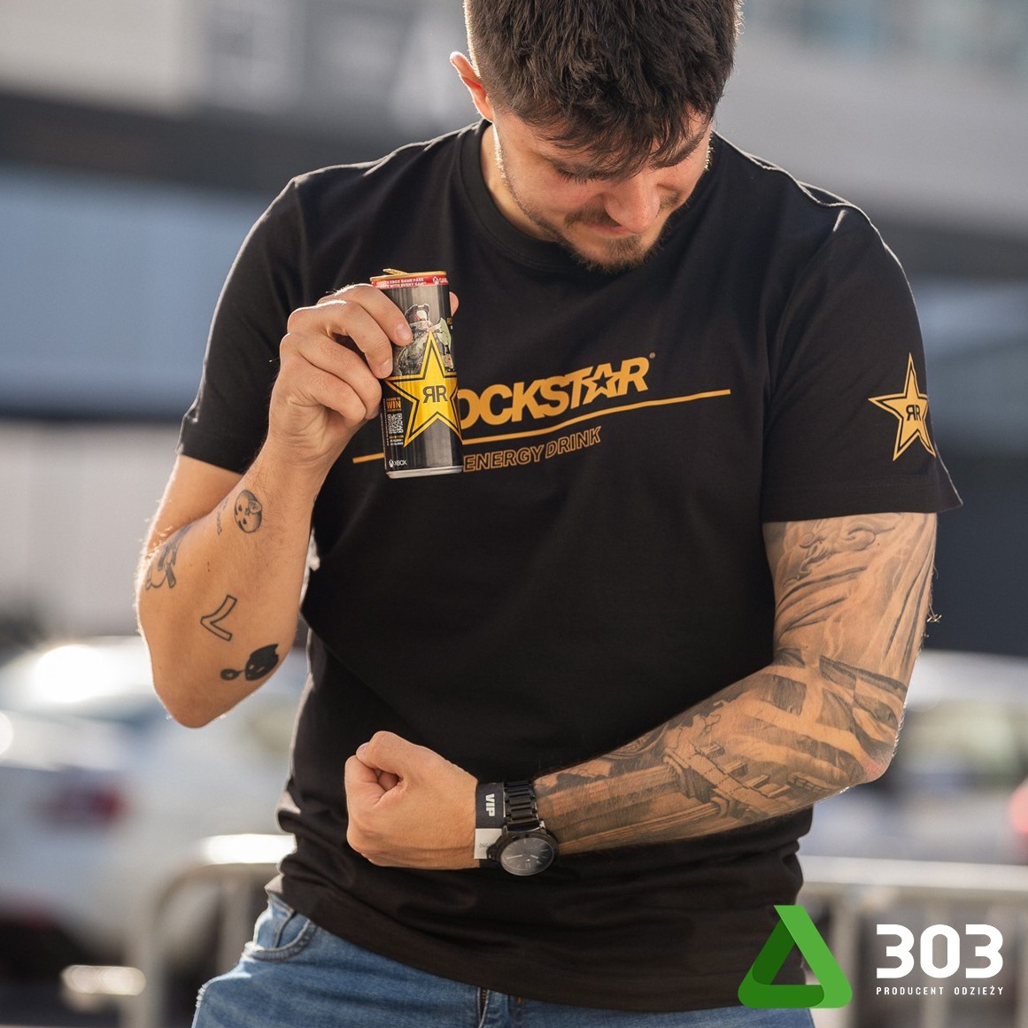 Mężczyzna w czarnej koszulce z logo Rockstar trzyma puszkę napoju energetycznego, prezentując nadruk. Widoczne tatuaże na rękach i zegarek. Ujęcie z bliska.