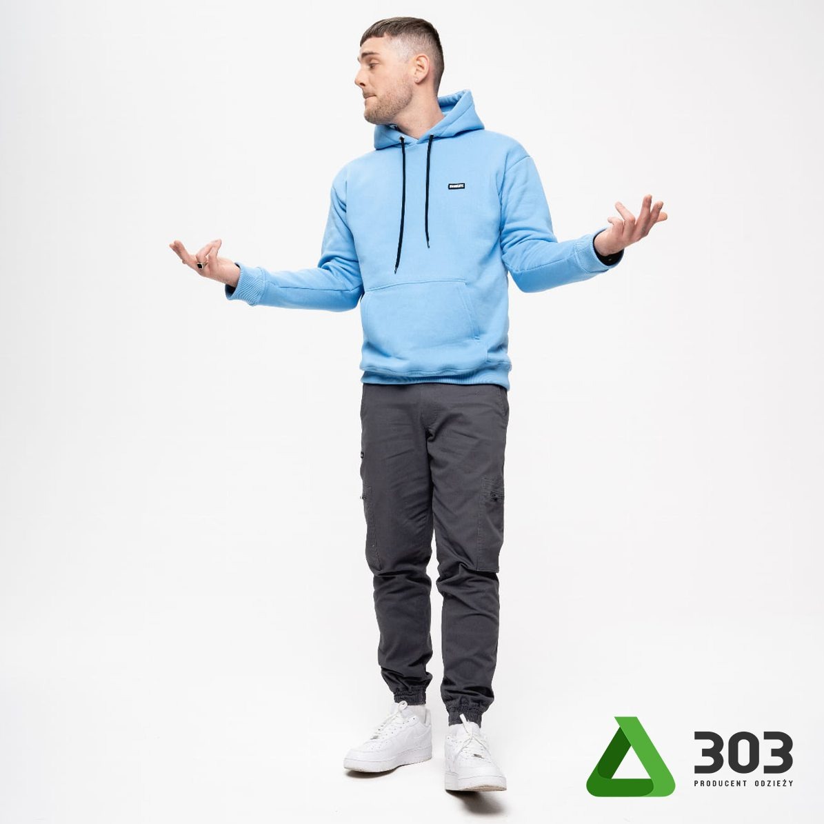 Mężczyzna w błękitnej bluzie z kapturem i szarych spodniach cargo, prezentujący produkt na białym tle z logo producenta odzieży w prawym dolnym rogu.