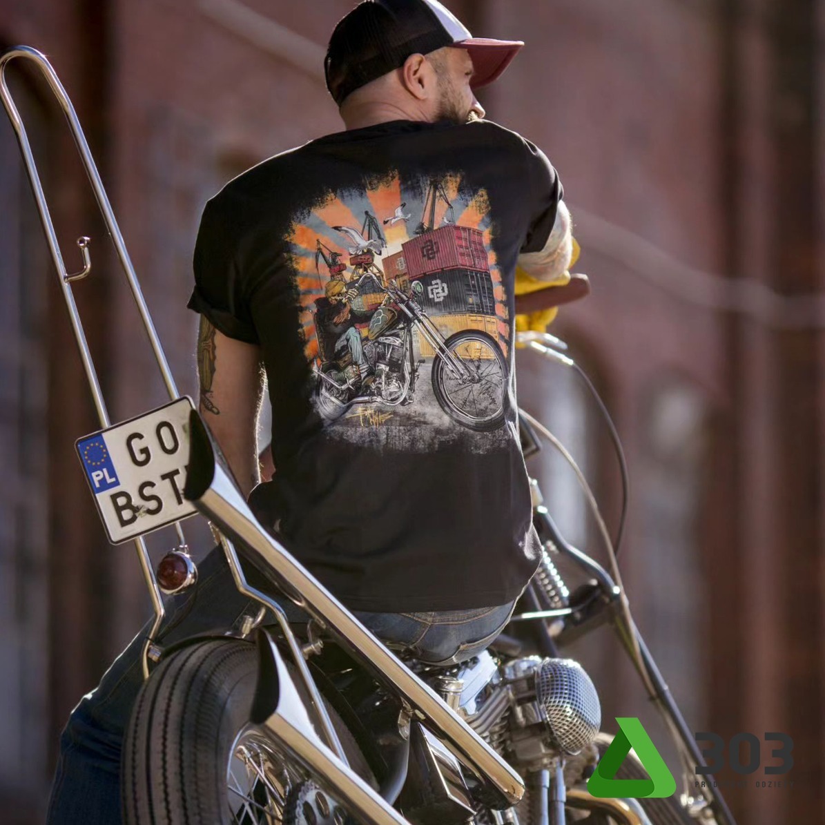 Mężczyzna w czarnej koszulce z nadrukiem motocykla i kontenerów, siedzący na chopperze. Widoczny tatuaż na ramieniu i tablica rejestracyjna. Styl vintage.